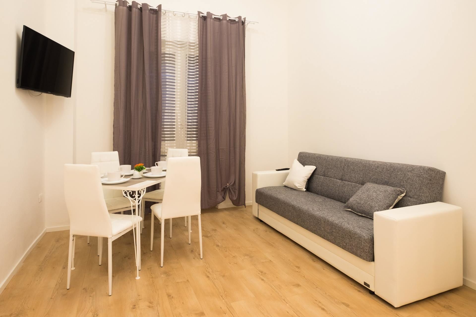 A small, modern living room with a grey sofa, white chairs, a small table with a TV, and a window with curtains.