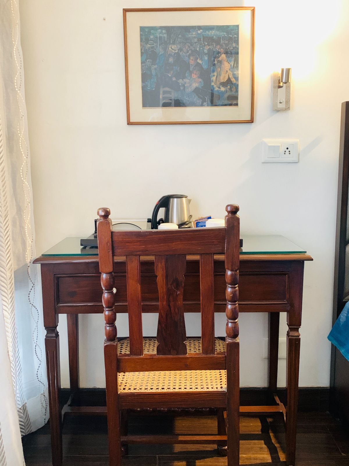 Une chaise en bois, dotée d’un siège et d’un dossier en osier, est placée devant un bureau sur lequel se trouvent une bouilloire et un tableau encadré accroché au mur.