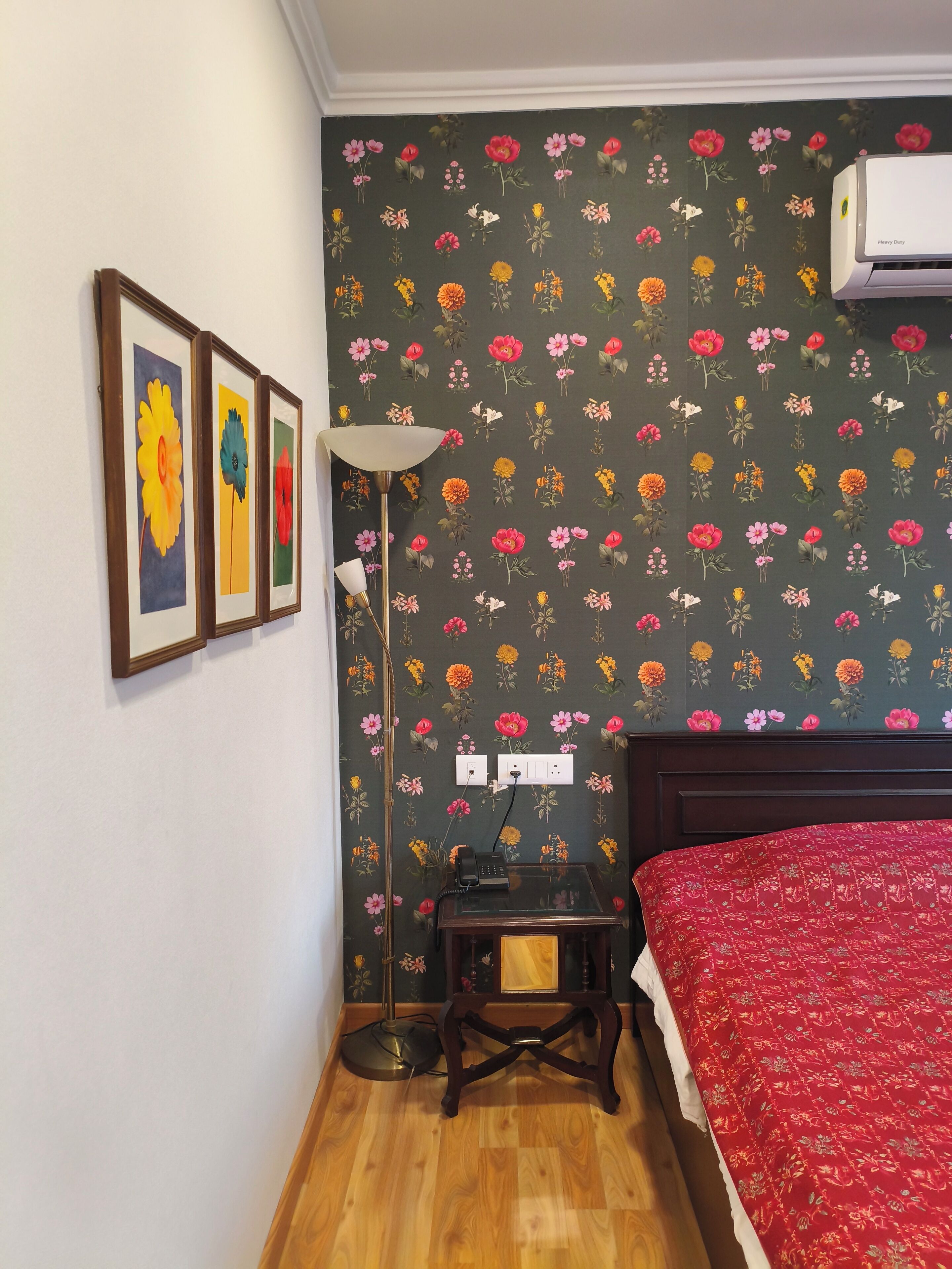 A bedroom with a floral wallpaper, a bed with a red cover, a bedside table with a lamp, and three framed pictures on the wall.