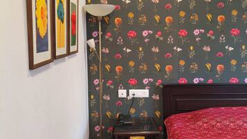 A bedroom with a floral wallpaper, a bed with a red cover, a bedside table with a lamp, and three framed pictures on the wall.