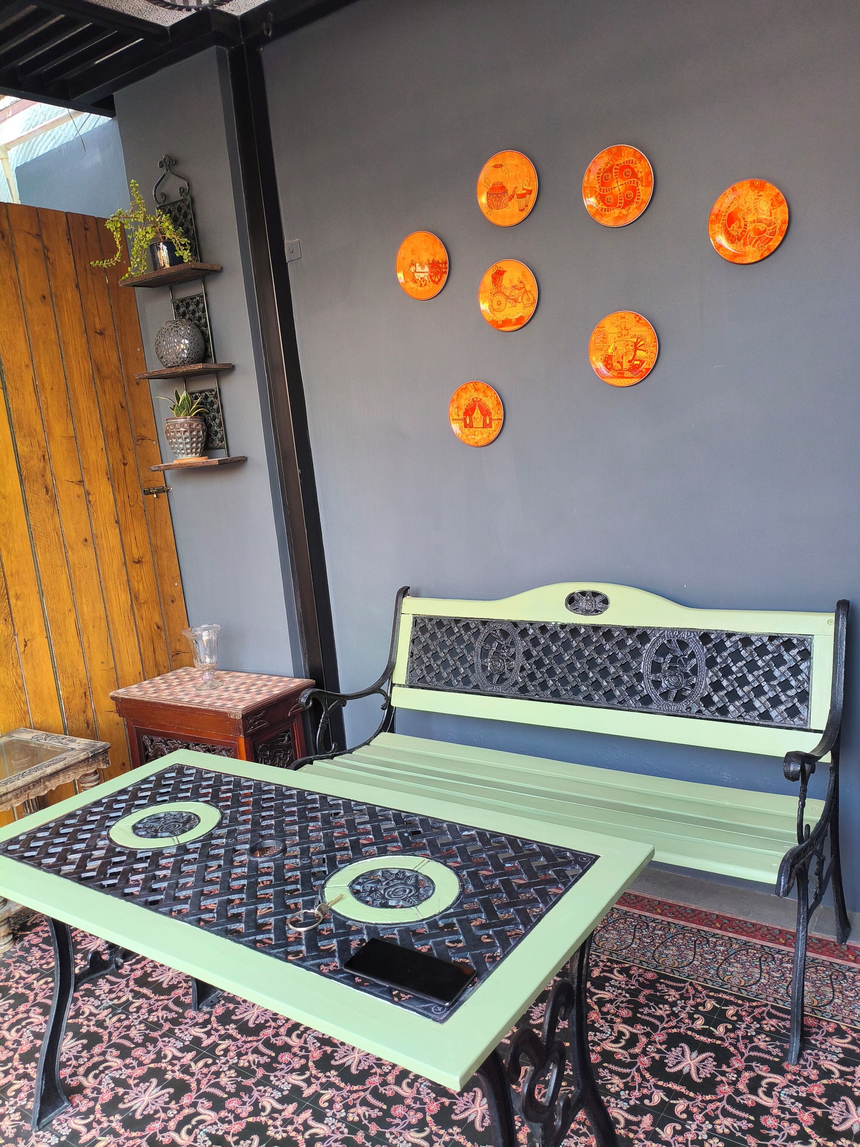 A cozy outdoor seating area with a green bench and a patterned table, surrounded by decorative plates on the wall.