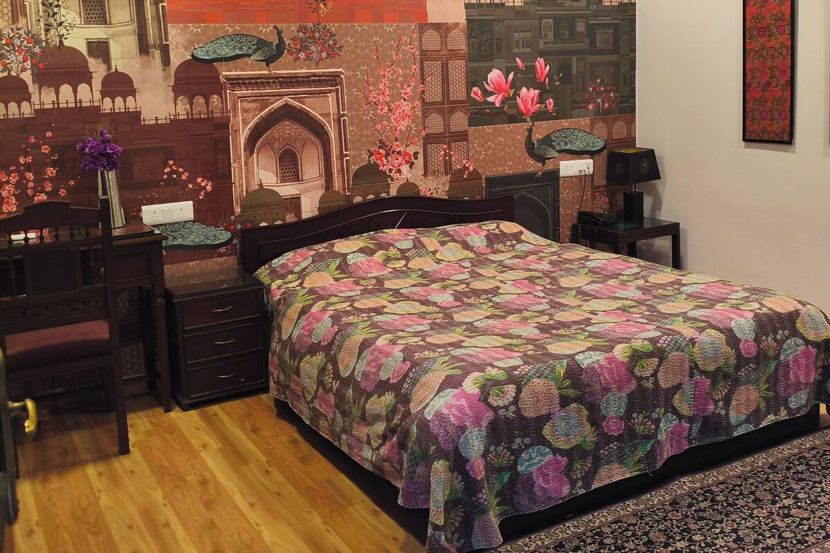 A bedroom with a large bed, a desk, a chair, a standing lamp, a wall-mounted air conditioner, and a ceiling fan.