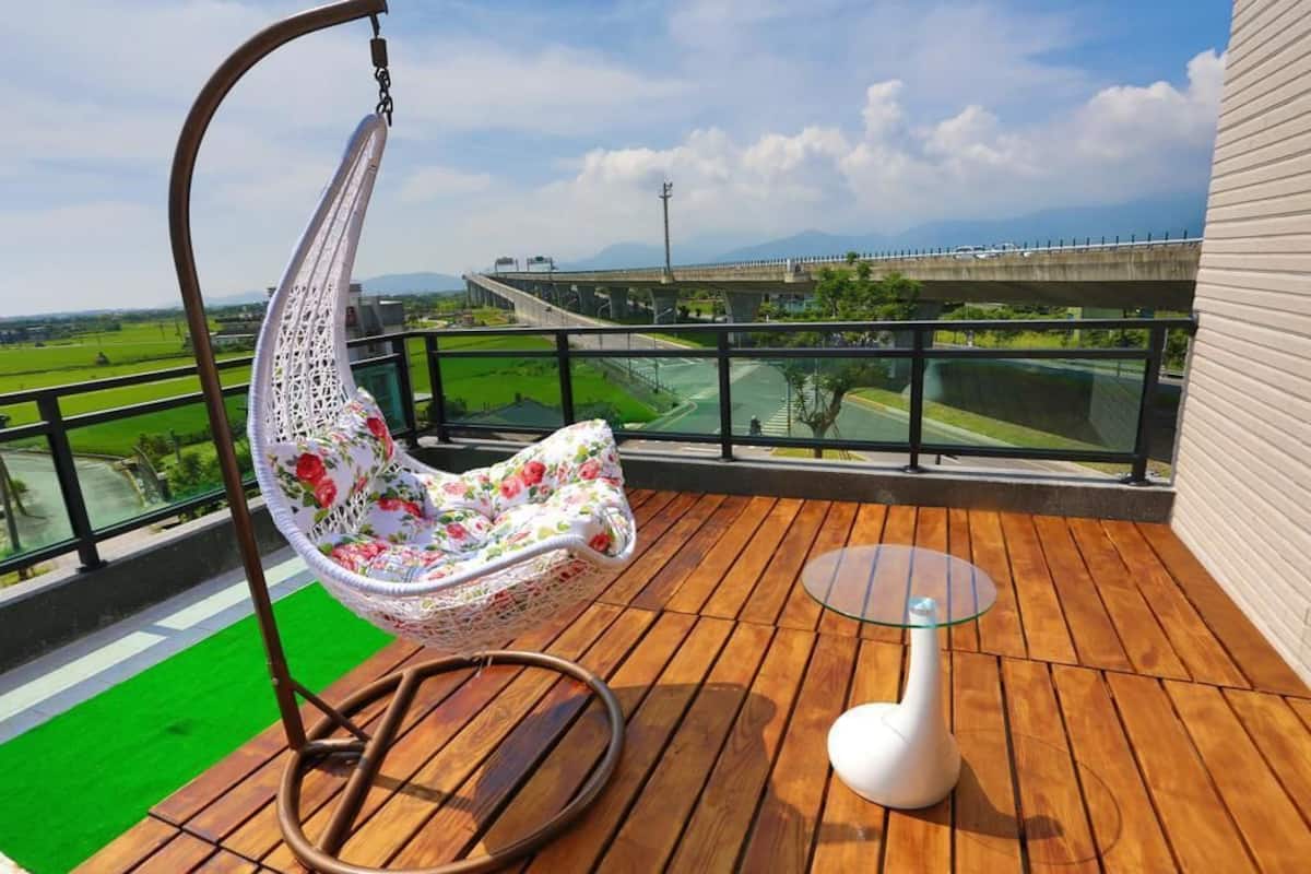A balcony with a hanging chair, a small table, and a view of a bridge and green fields.