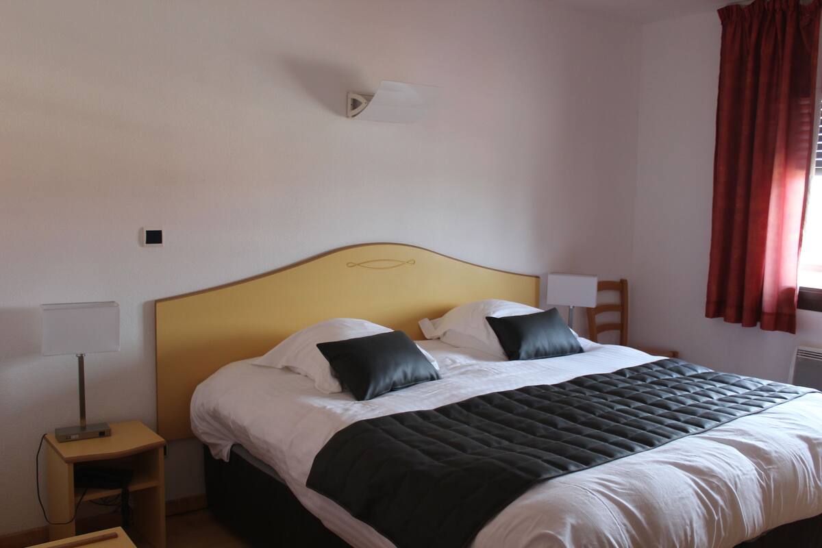 Una cama bien tendida con cabecera, una mesita de noche con lámpara y una ventana con cortinas.