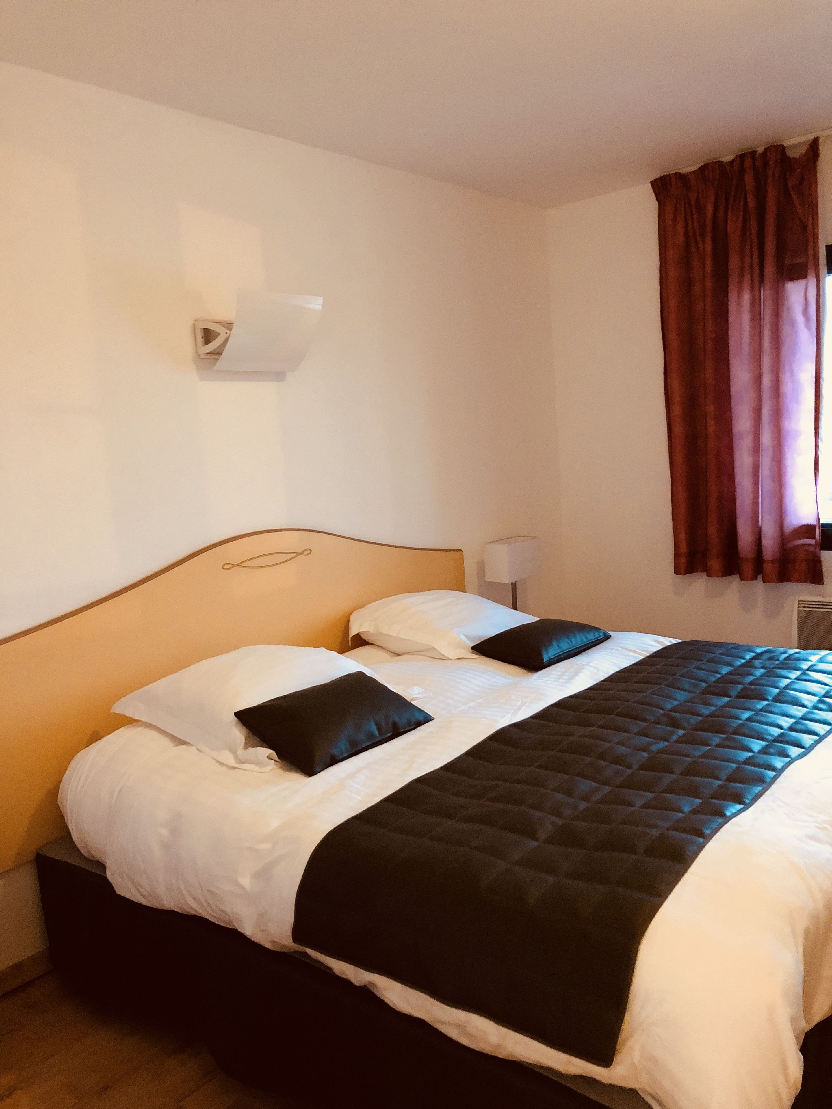 Une chambre d’hôtel avec un lit, du linge de lit blanc, une couverture noire matelassée et un rideau marron.