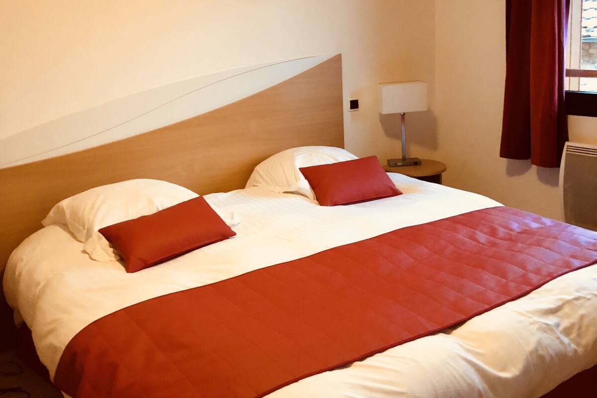Habitación de hotel con una cama grande, ropa de cama roja y blanca, una lámpara de noche y una ventana con cortinas.