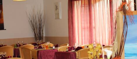 A dining area with tables set for a meal, featuring a view of the sea through a window with sheer curtains.