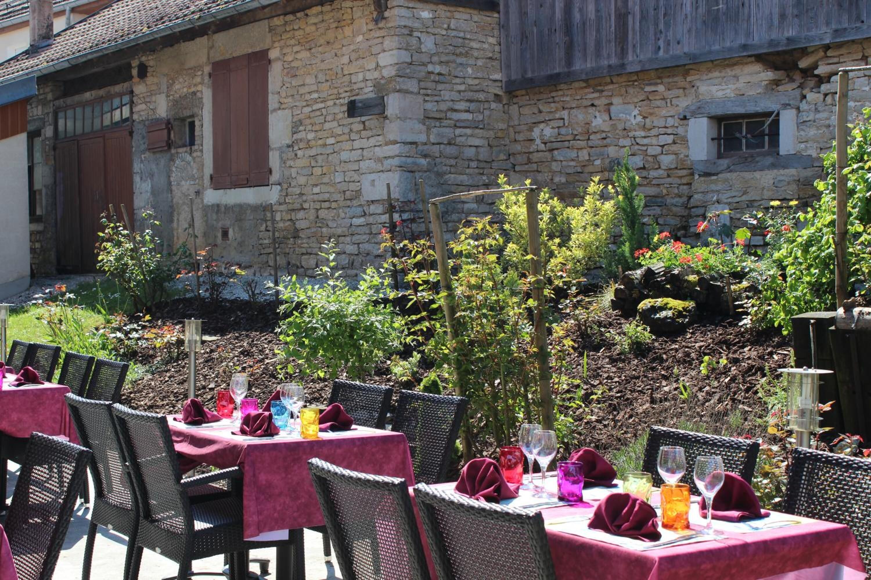 Un área de comedor al aire libre con mesas dispuestas para dos, rodeada de muros de piedra y follaje.