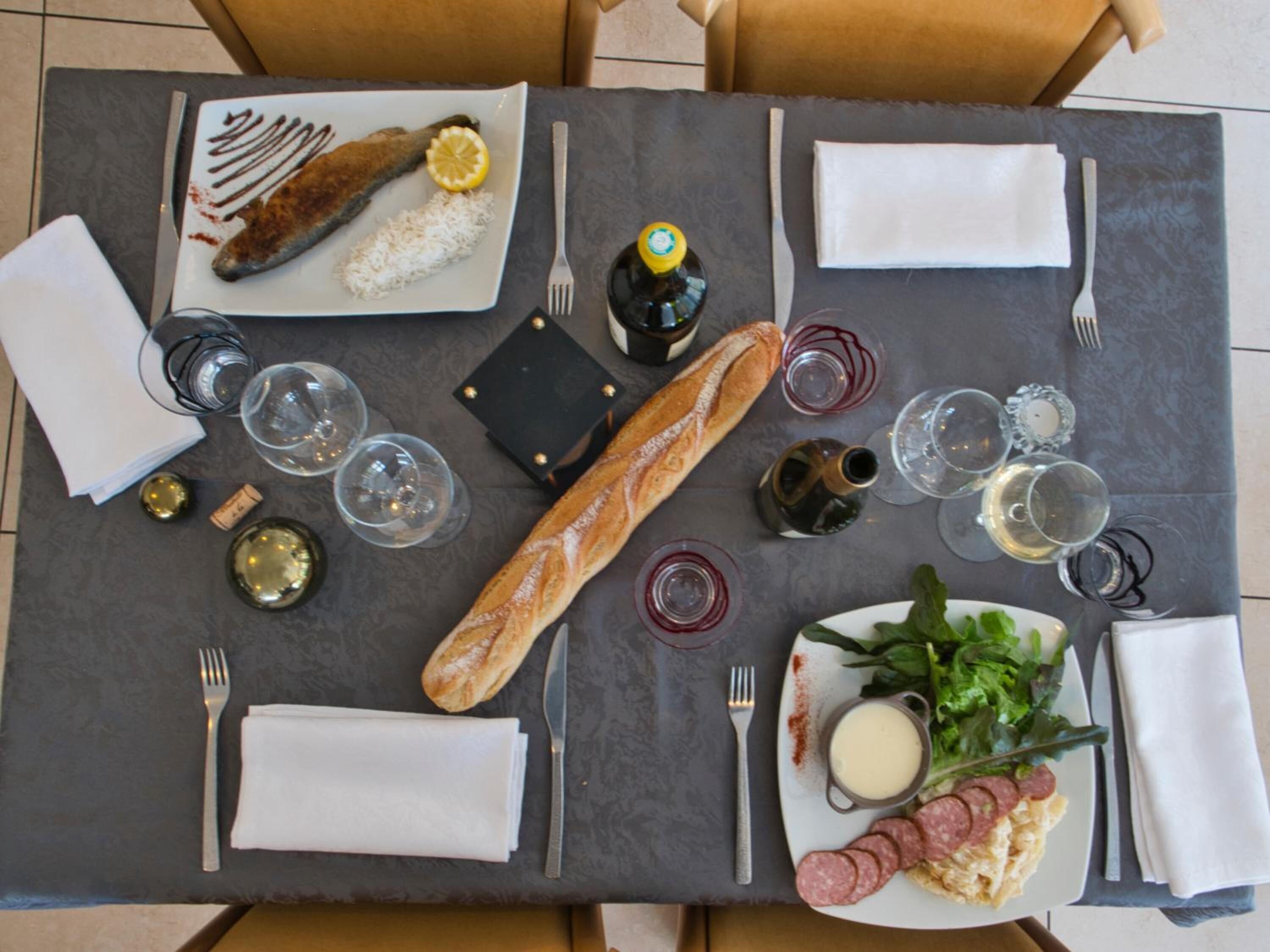 Una mesa de comedor servida con un plato de pescado, pan, copas de vino y un plato con ensalada y salami.