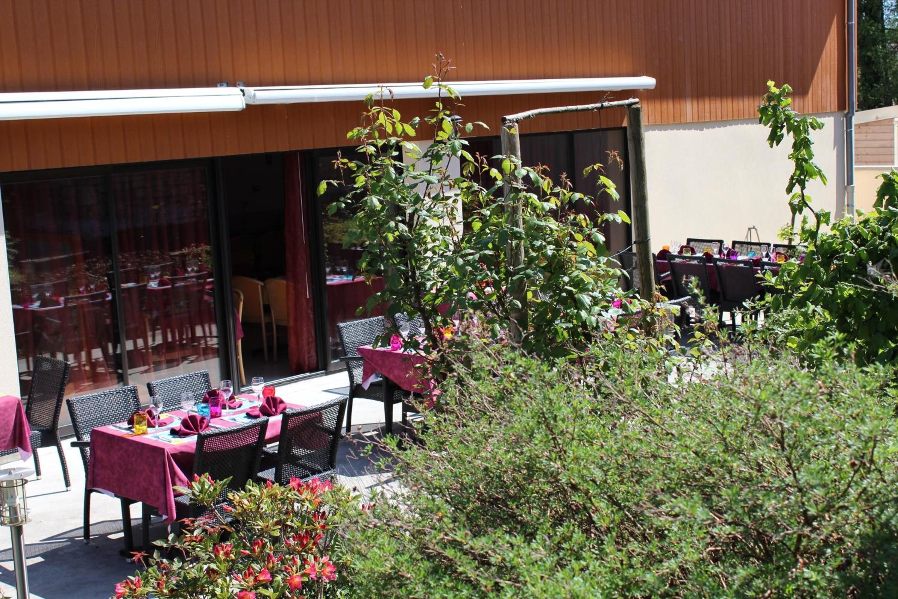 Un espace de restauration en plein air, avec des tables et des chaises, entouré de verdure et bordé d’un bâtiment doté de grandes fenêtres.