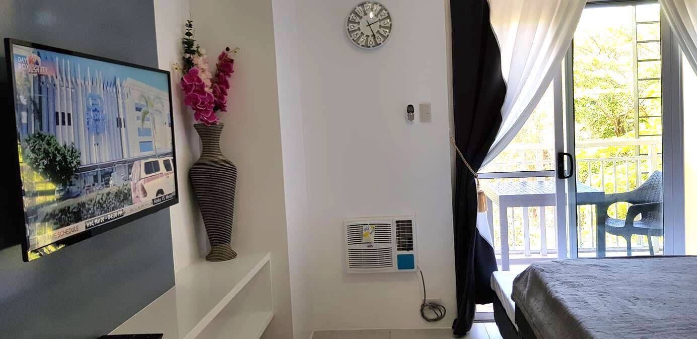 A room with a TV displaying a game schedule, a large vase with flowers, a clock, and a window with curtains.