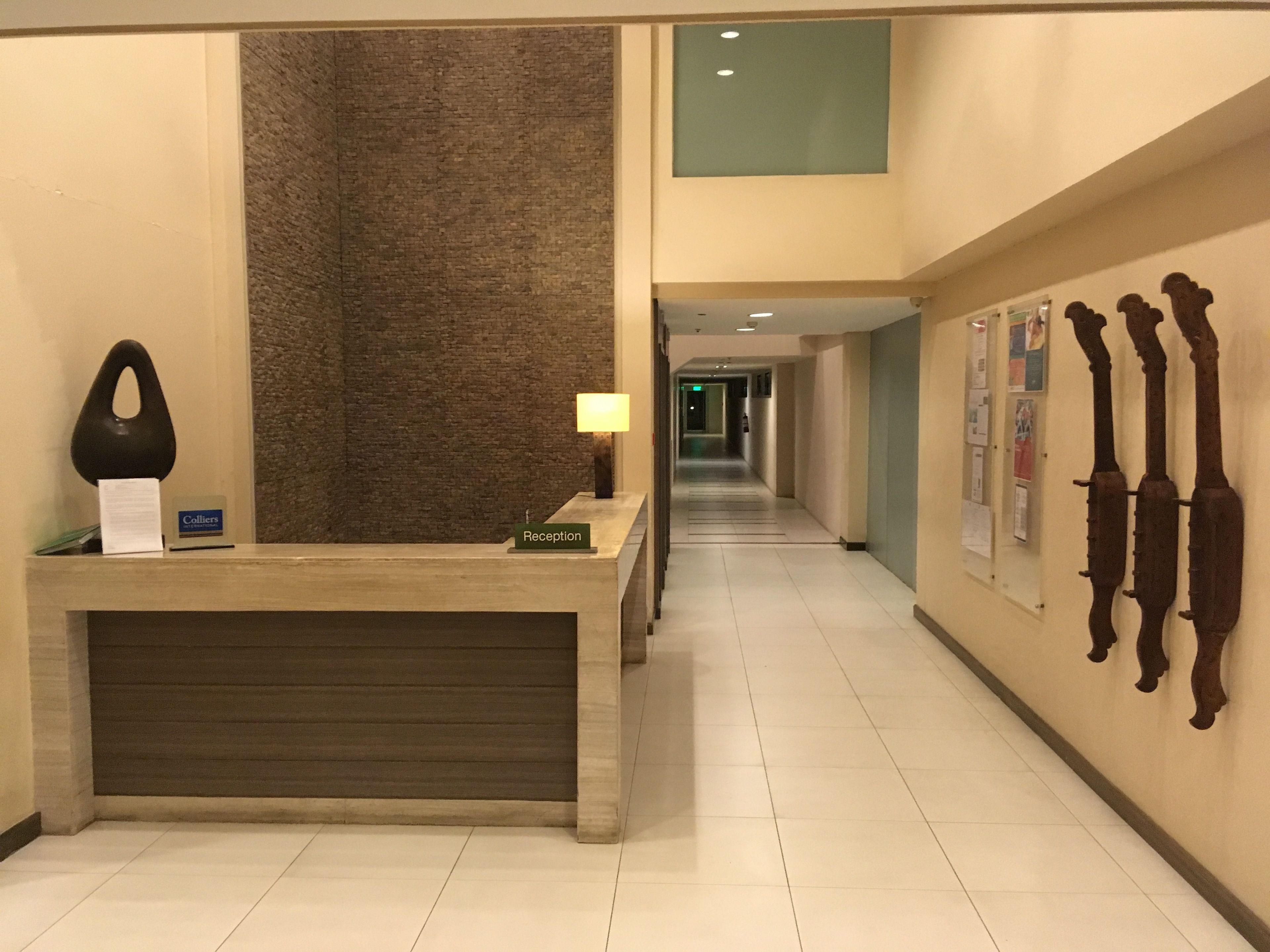 A hotel reception area with a reception desk, a lamp, a sign indicating the reception, and a corridor leading to other parts of the hotel.