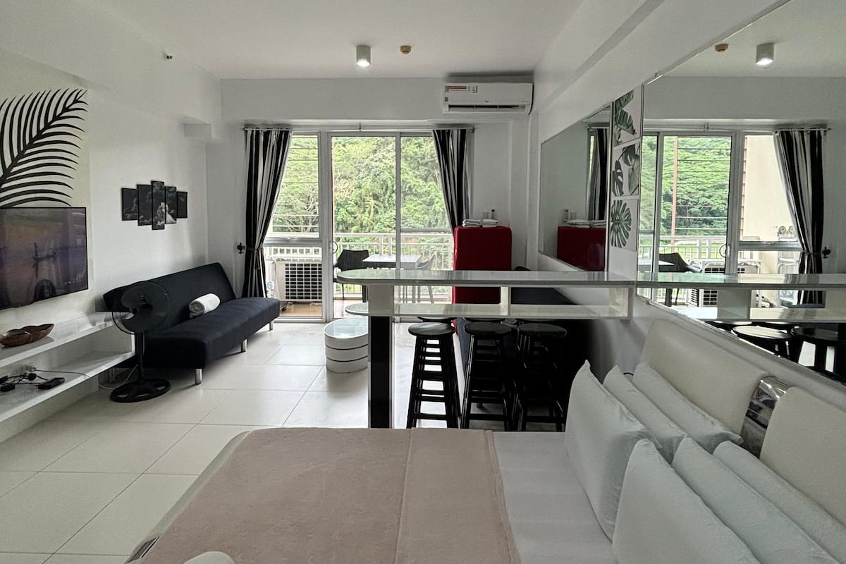 A modern hotel room with a large bed, a sofa, a dining table, and a balcony with a view.