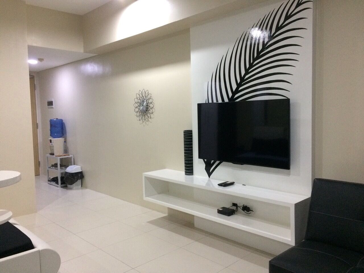 A modern living room with a flat-screen TV mounted on a white console, a decorative wall piece, and a water dispenser in the background.