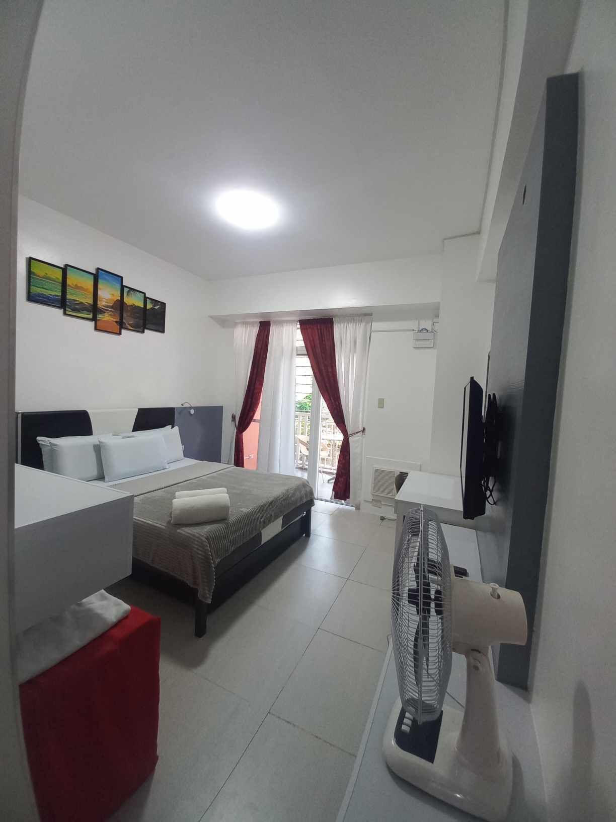 Habitació d'hotel amb llit, escriptori, cadira, ventilador i finestra amb cortines.