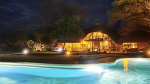 A well-lit resort with a swimming pool, a large tree, and a building with a thatched roof. - Tawi Lodge (Amboseli)