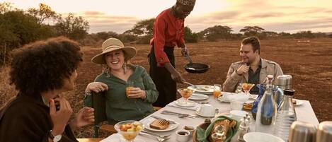 A group of people dining outdoors at a table set with various dishes, utensils, and a bottle.