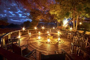 An outdoor circular seating area with a fire pit at the center, surrounded by chairs and tables, under a night sky with visible clouds. - Tawi Lodge (Amboseli)