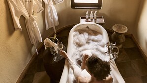 A bathroom with a bathtub filled with bubbles, a wine bottle, and glasses on a small table. There are two white robes hanging on hooks on the wall. - Tawi Lodge (Amboseli)