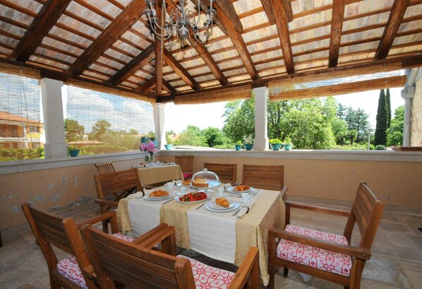 Un área de comedor al aire libre techada, con una mesa dispuesta para una comida, rodeada de sillas de madera con cojines estampados.