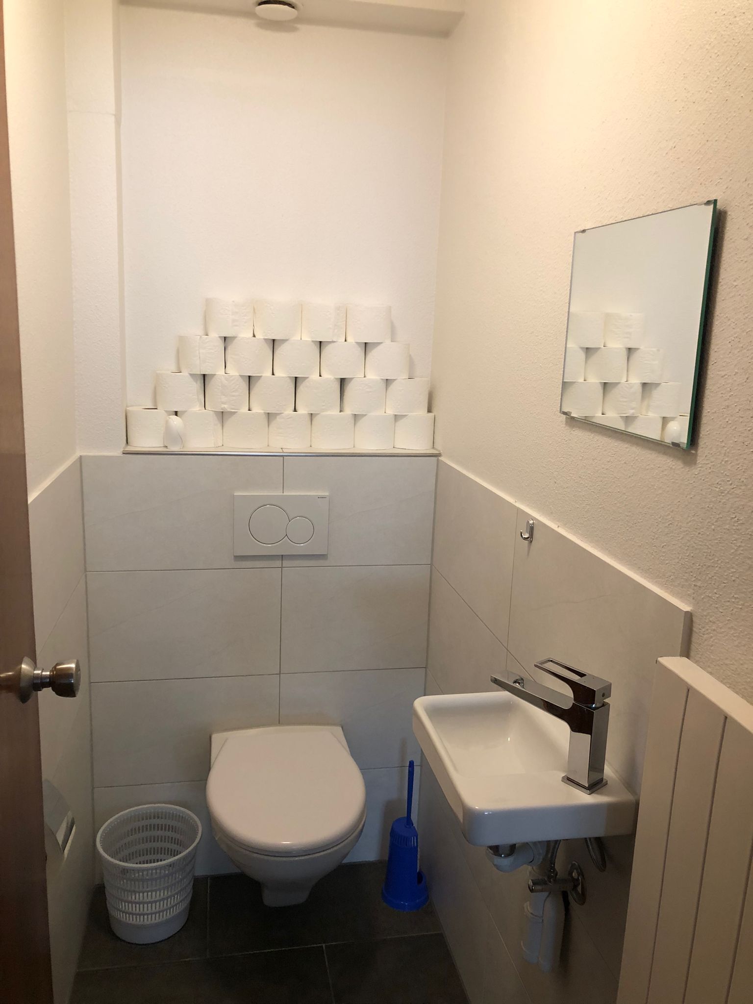 Une petite salle de bain propre, équipée de toilettes, d’un lavabo et d’un porte-rouleau de papier toilette fixé au mur.