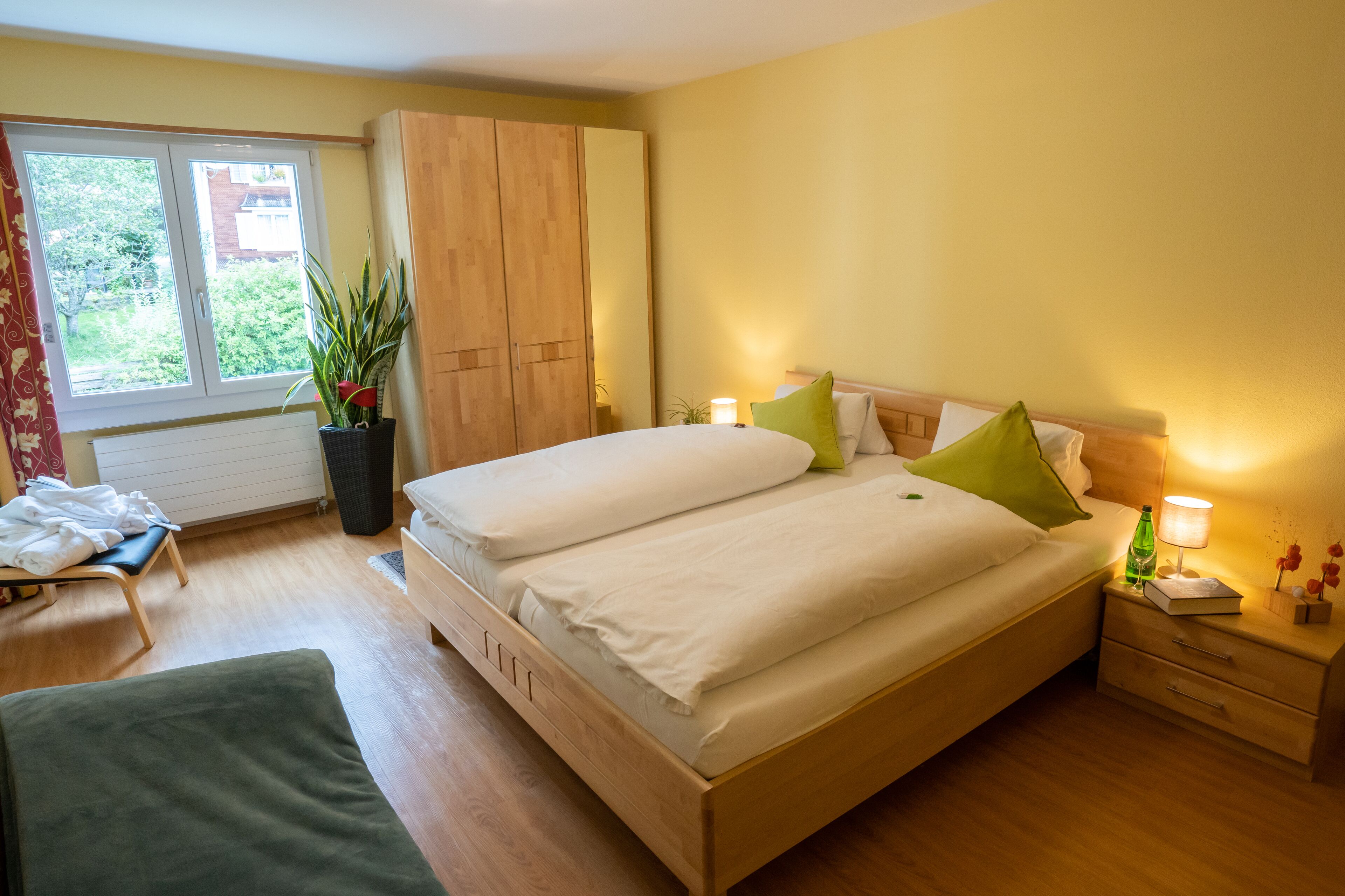 Un dormitorio con una cama doble, mesitas de noche, un armario, un ventanal con cortinas y una planta en maceta.