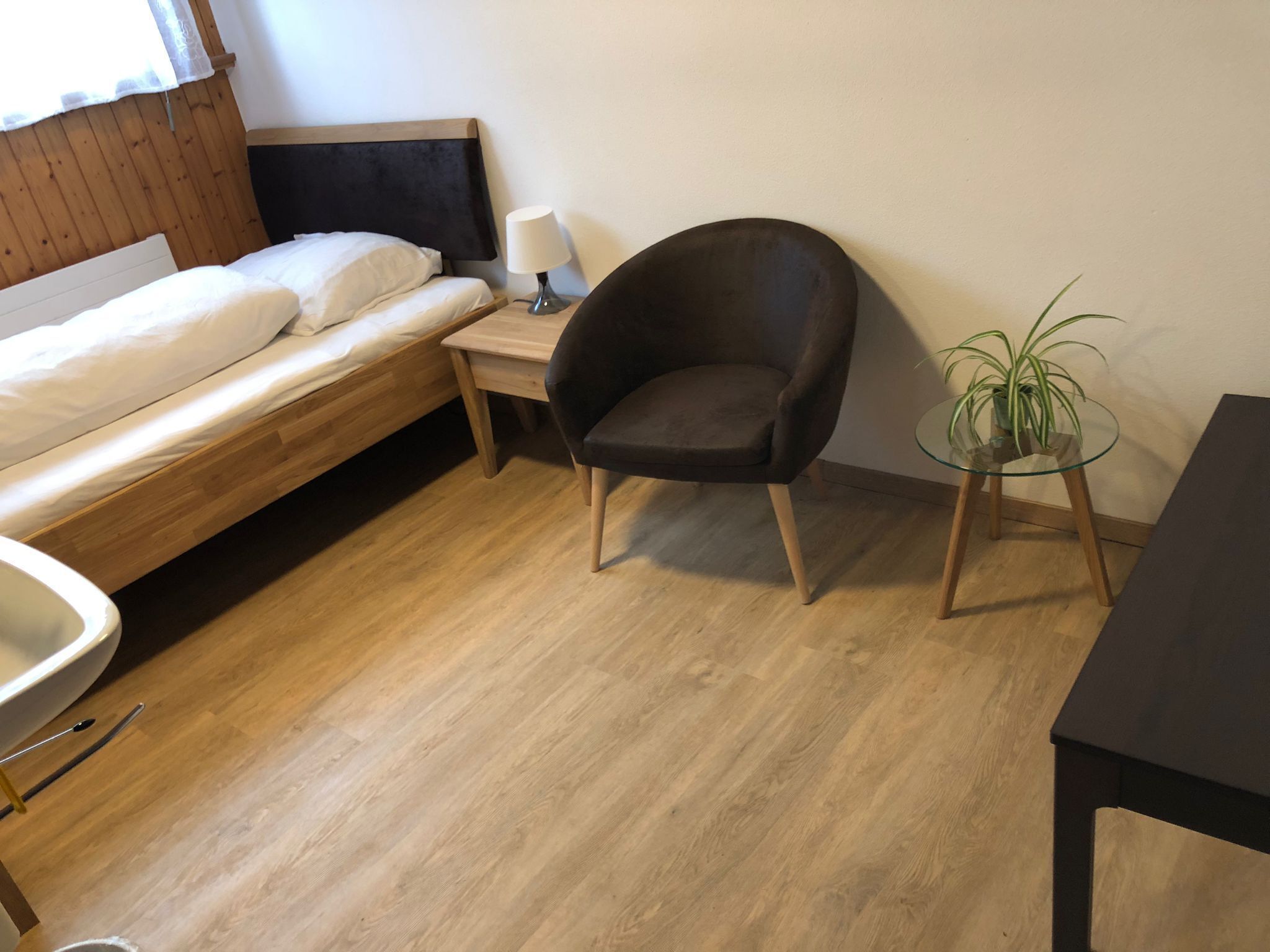 Una habitación pequeña y ordenada con una cama, una silla, una mesita de noche con una lámpara, una mesa redonda pequeña con una planta y un espejo grande.