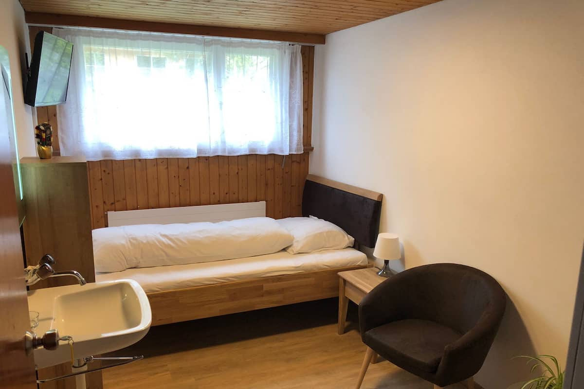 Une petite pièce agréable et chaleureuse, avec un lit en bois, une table de chevet avec une lampe, une chaise et un lavabo.