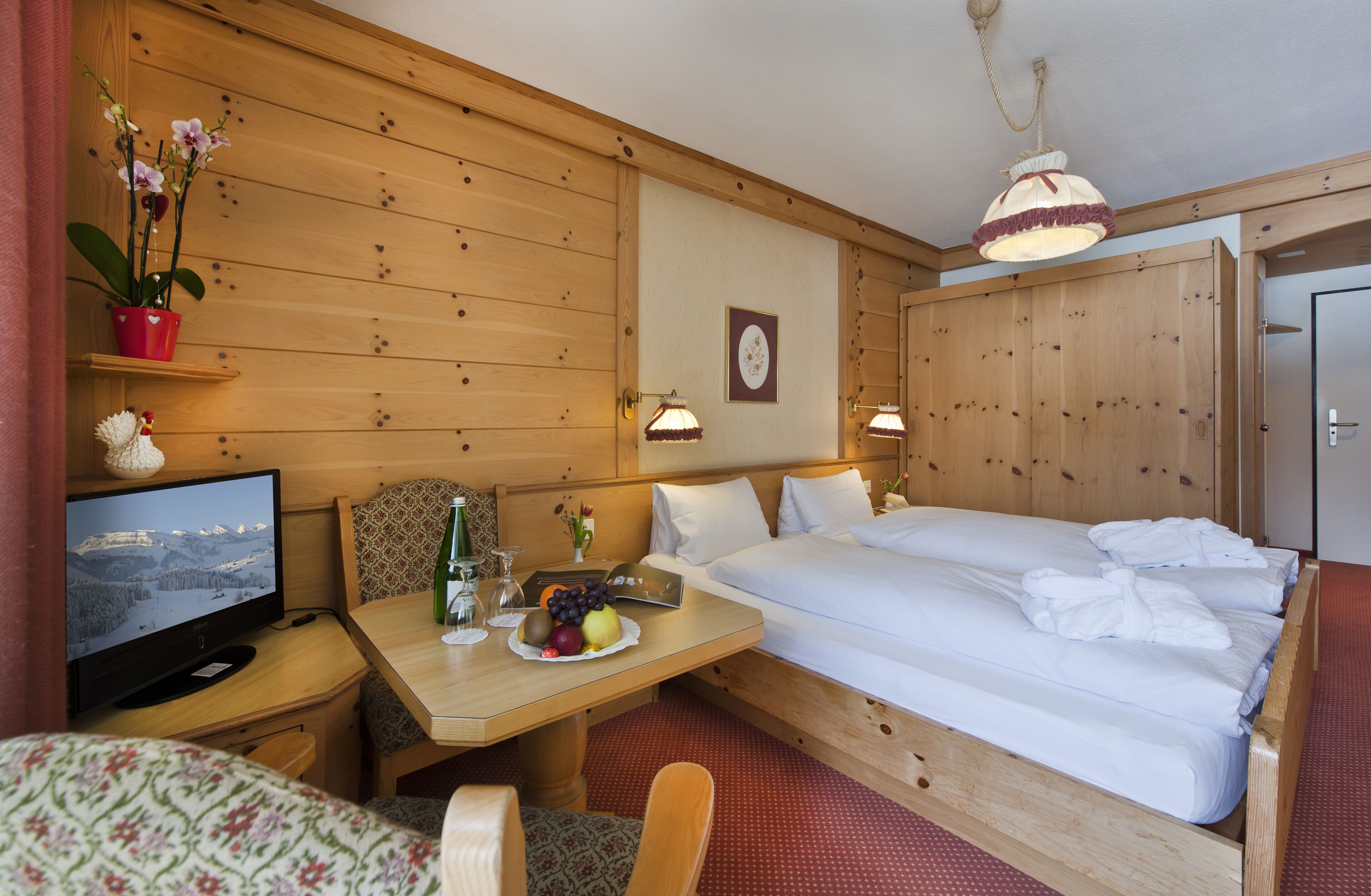 Habitación de hotel con decoración de madera, una cama, un televisor, una mesa con fruta, una planta y un cuadro colgado en la pared.