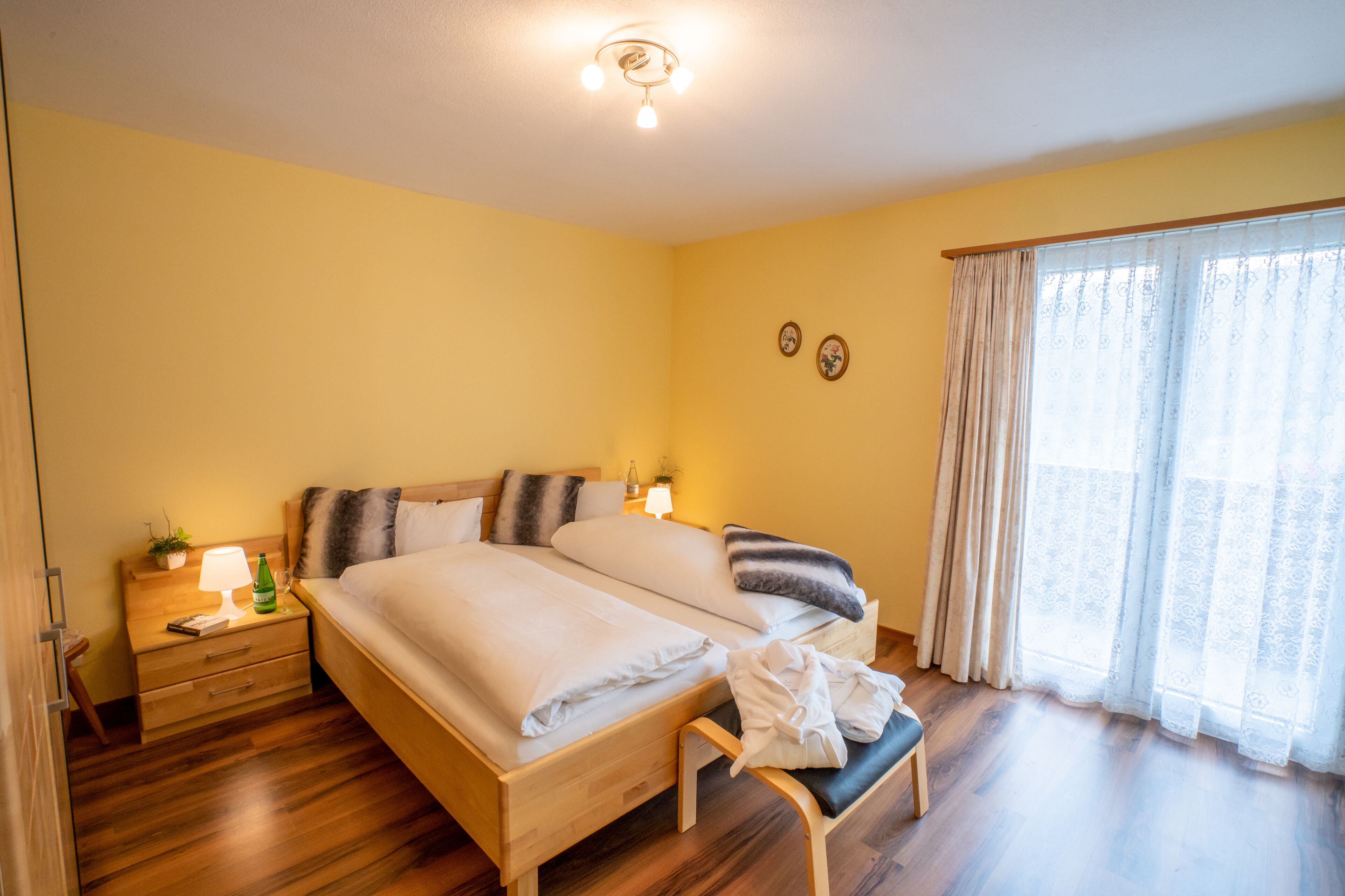 Habitación de hotel con dos camas, piso de madera, una mesita de noche con lámpara y una ventana con cortinas.