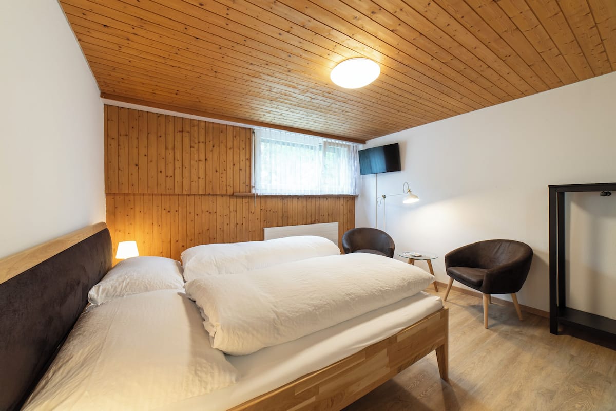 Une chambre avec un plafond en bois, un lit recouvert de draps blancs, une petite table avec une lampe et une chaise.