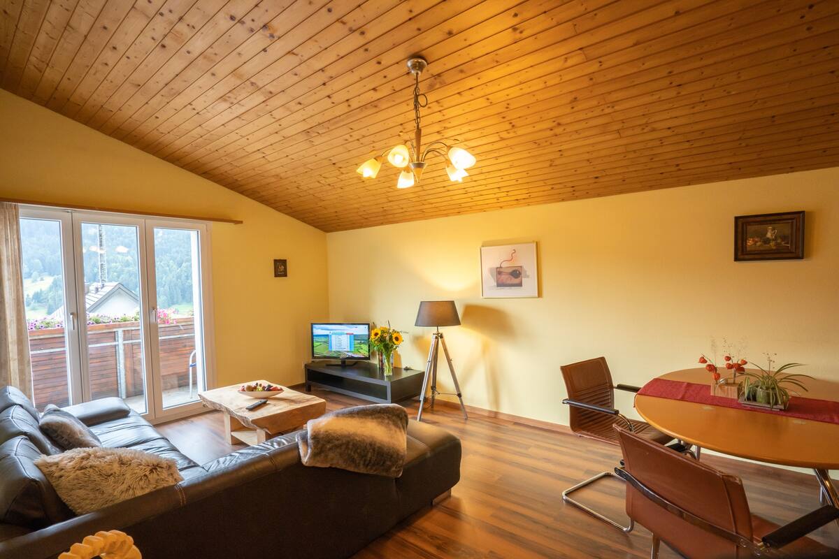 Un salon avec un plafond en bois, un canapé, un téléviseur, une table à manger avec des chaises et une fenêtre donnant sur un jardin.