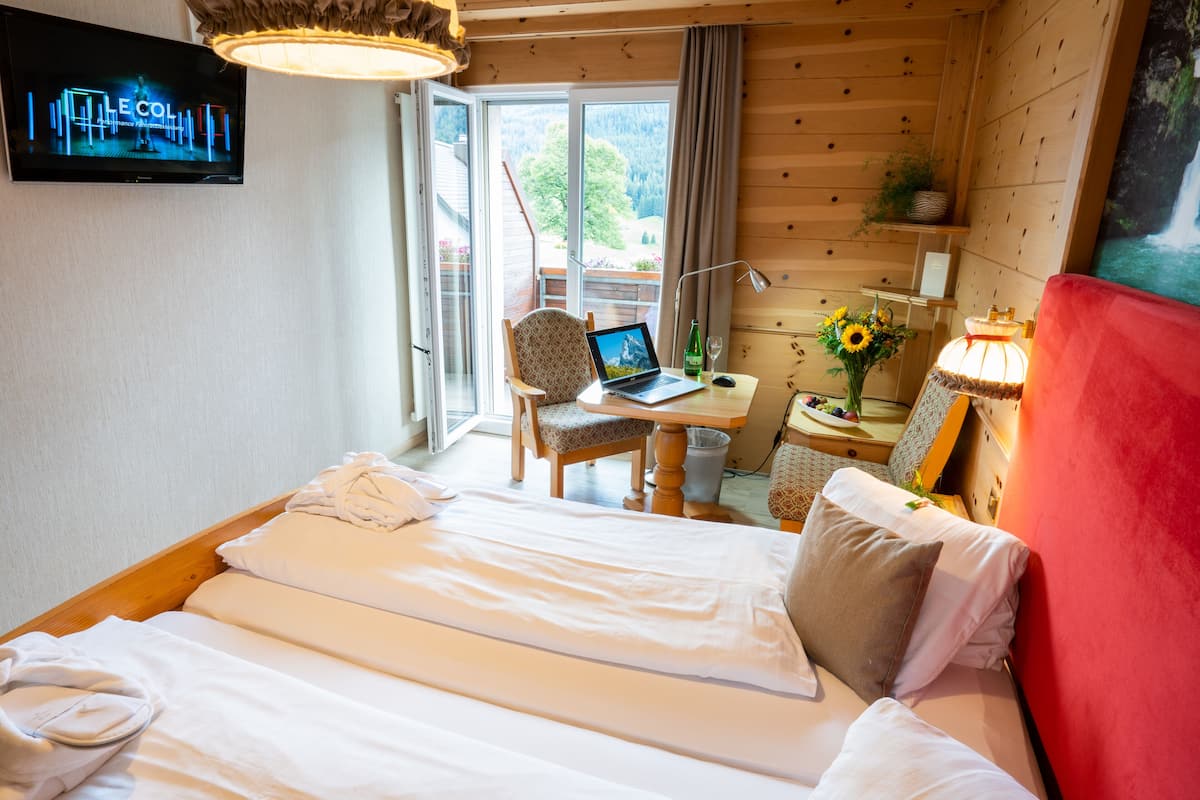 Une chambre d’hôtel avec un lit, un bureau avec un ordinateur portable, une télévision et une fenêtre donnant sur des arbres.