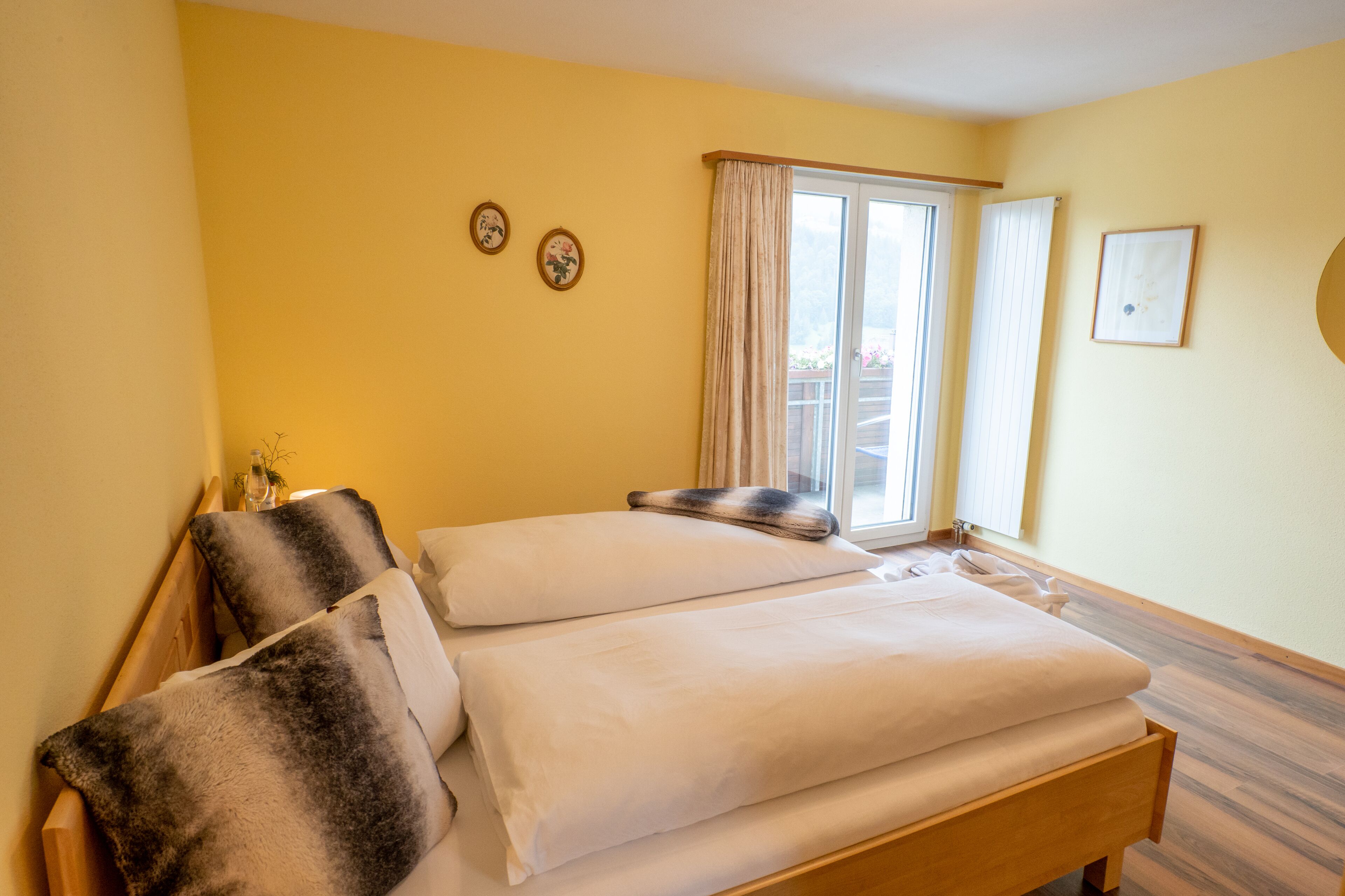 A double bed with white and dark gray linens, a wooden headboard, and a window with curtains.