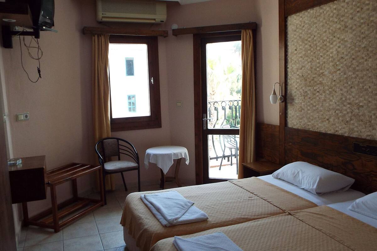 A hotel room with two beds, a small table with a chair, a window with curtains, and a balcony with chairs.
