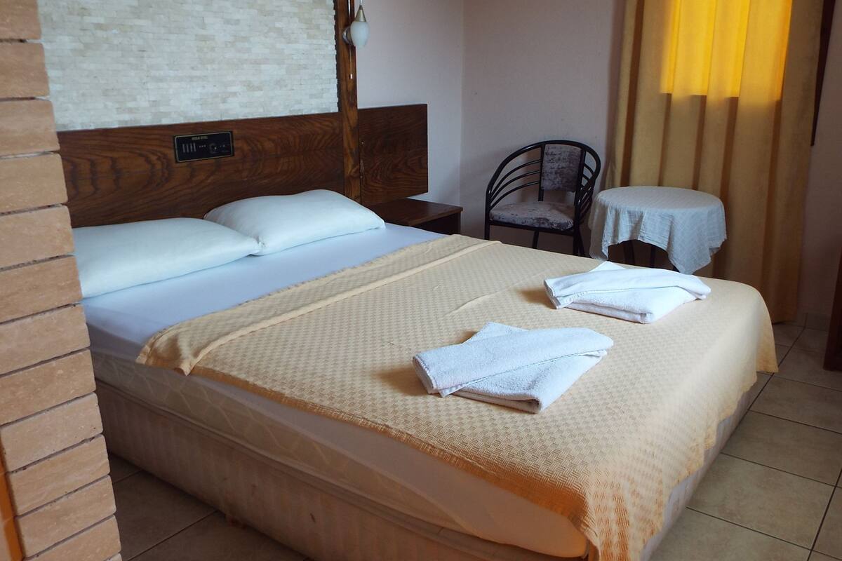 A neatly made bed with a headboard, a small round table with a tablecloth, a chair, and a bedside table with a lamp.