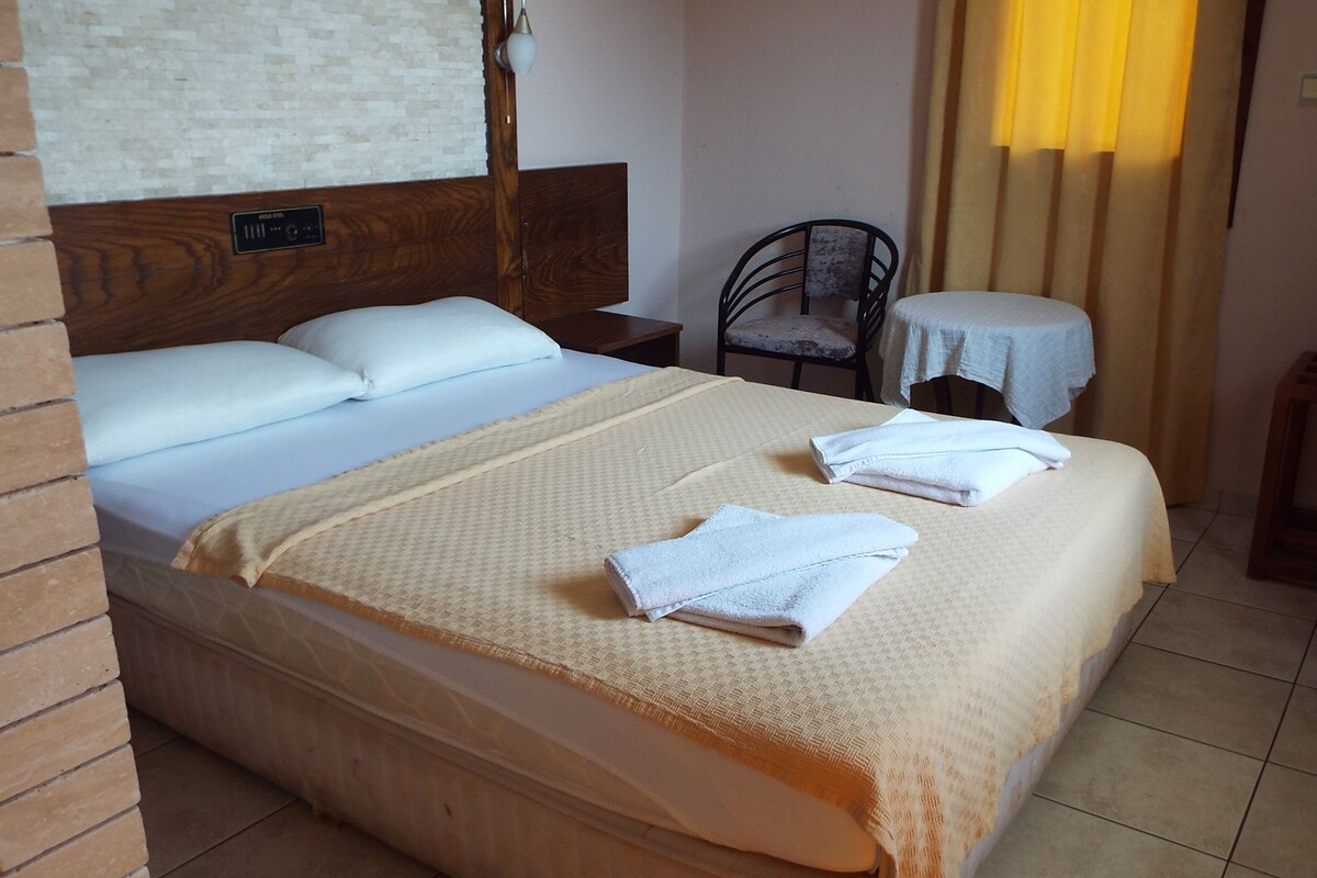 A neatly made bed with pillows and folded towels, a wooden headboard, a small round table with a chair, and a window with curtains.