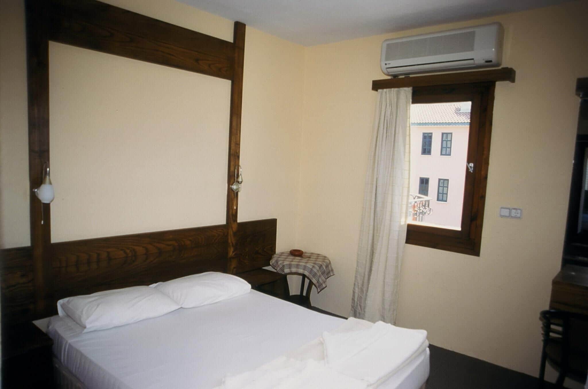A hotel room with a bed, a small table with a tablecloth, an air conditioning unit, and a window with a view of buildings.