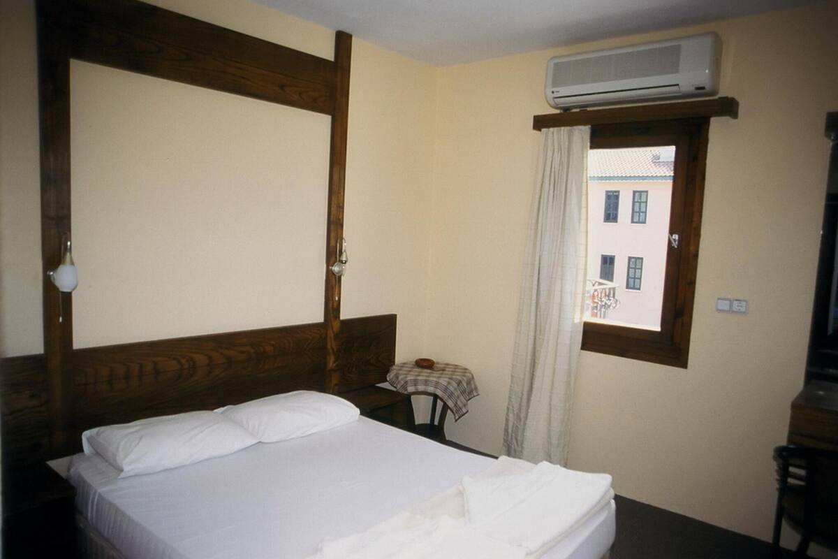 A hotel room with a bed, a small table with a lamp, a window with curtains, and an air conditioning unit.