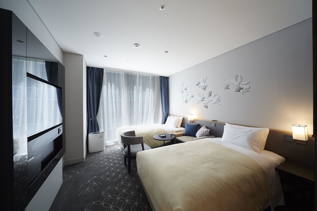 Hotelkamer met een bed, nachtkastjes, een bureau, een stoel, een televisie en een groot raam met gordijnen.