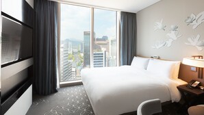 A hotel room with a large bed, a desk with a lamp, a TV, and a view of the cityscape through the window. - Hotel Gracery Seoul (Seoul)