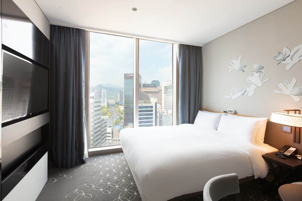 A hotel room with a large bed, a desk with a lamp, a TV, and a view of the cityscape through the window.