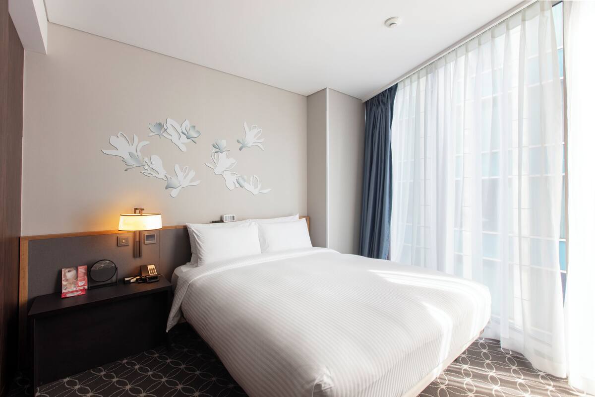 A hotel room with a bed, a nightstand with a lamp, a wall-mounted phone, and a window with sheer curtains.