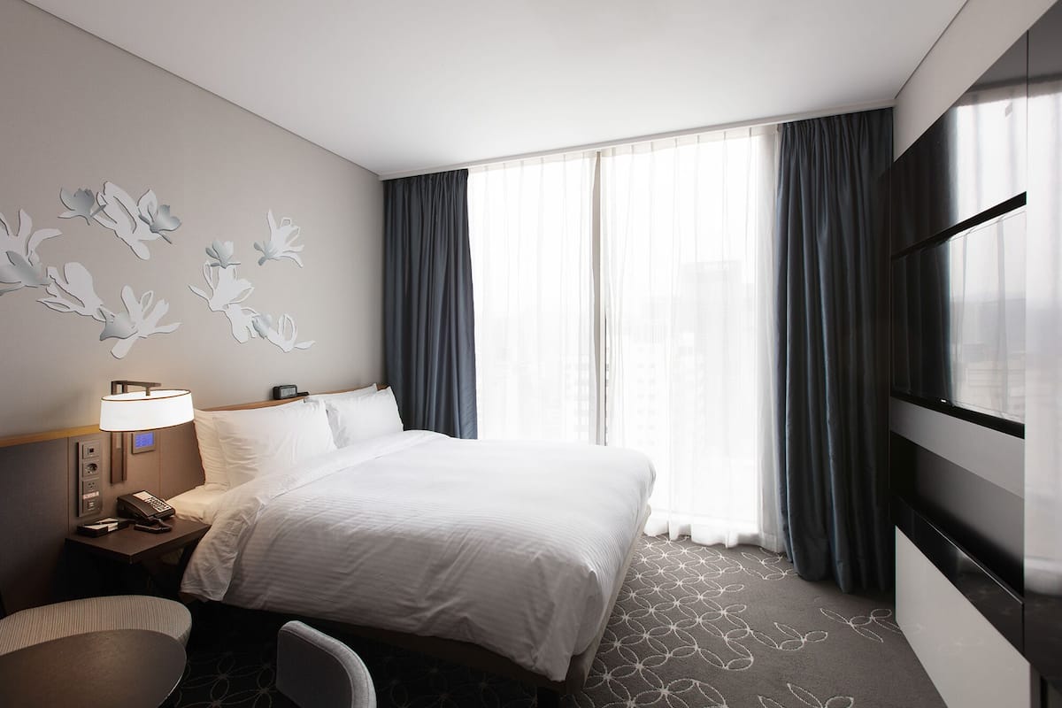 Une chambre d’hôtel avec un lit, un bureau avec une lampe, une chaise, une télévision et une fenêtre avec des rideaux.
