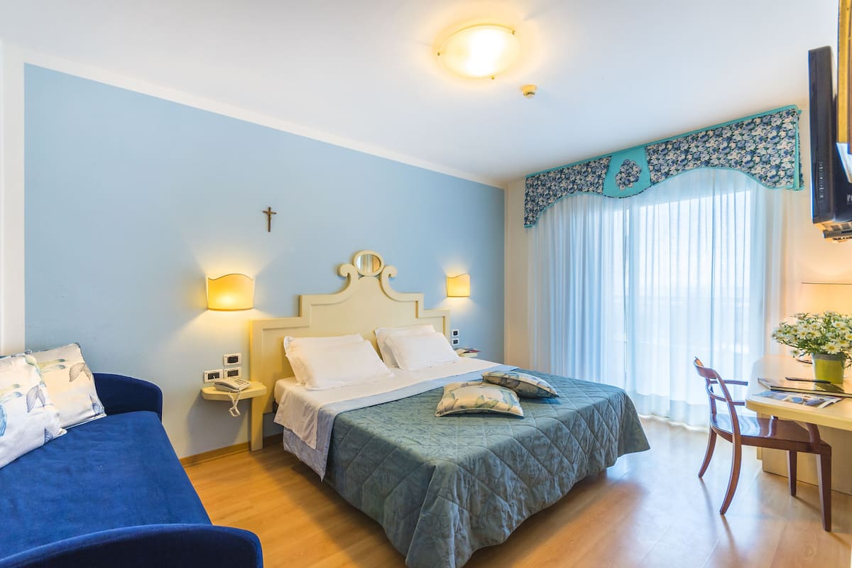 A hotel room with a bed, a desk, a chair, a small table, and a window with curtains.