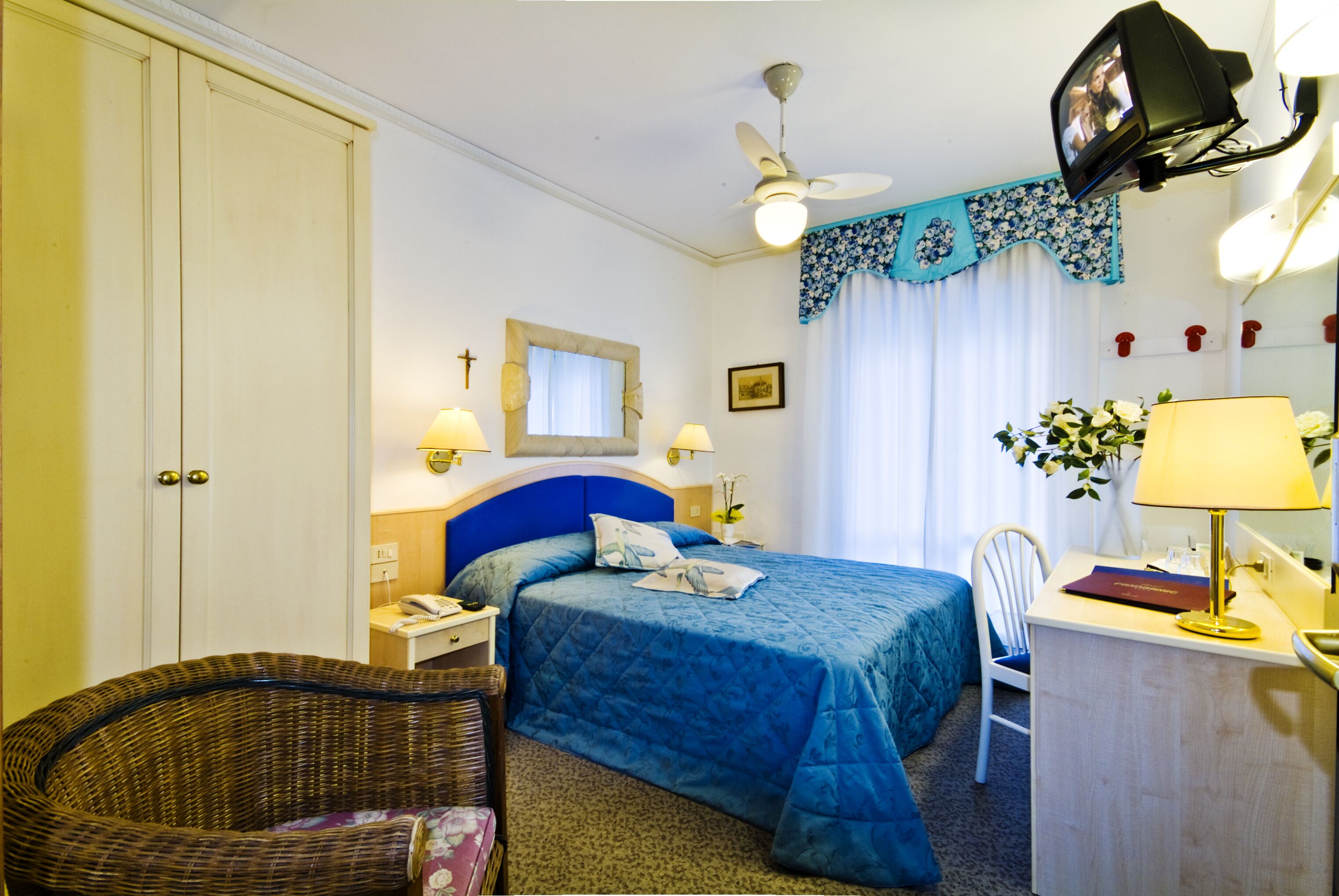 Une chambre d’hôtel équipée d’un lit, d’un bureau, d’une chaise, d’une télévision, d’un ventilateur de plafond et d’une fenêtre avec des rideaux.