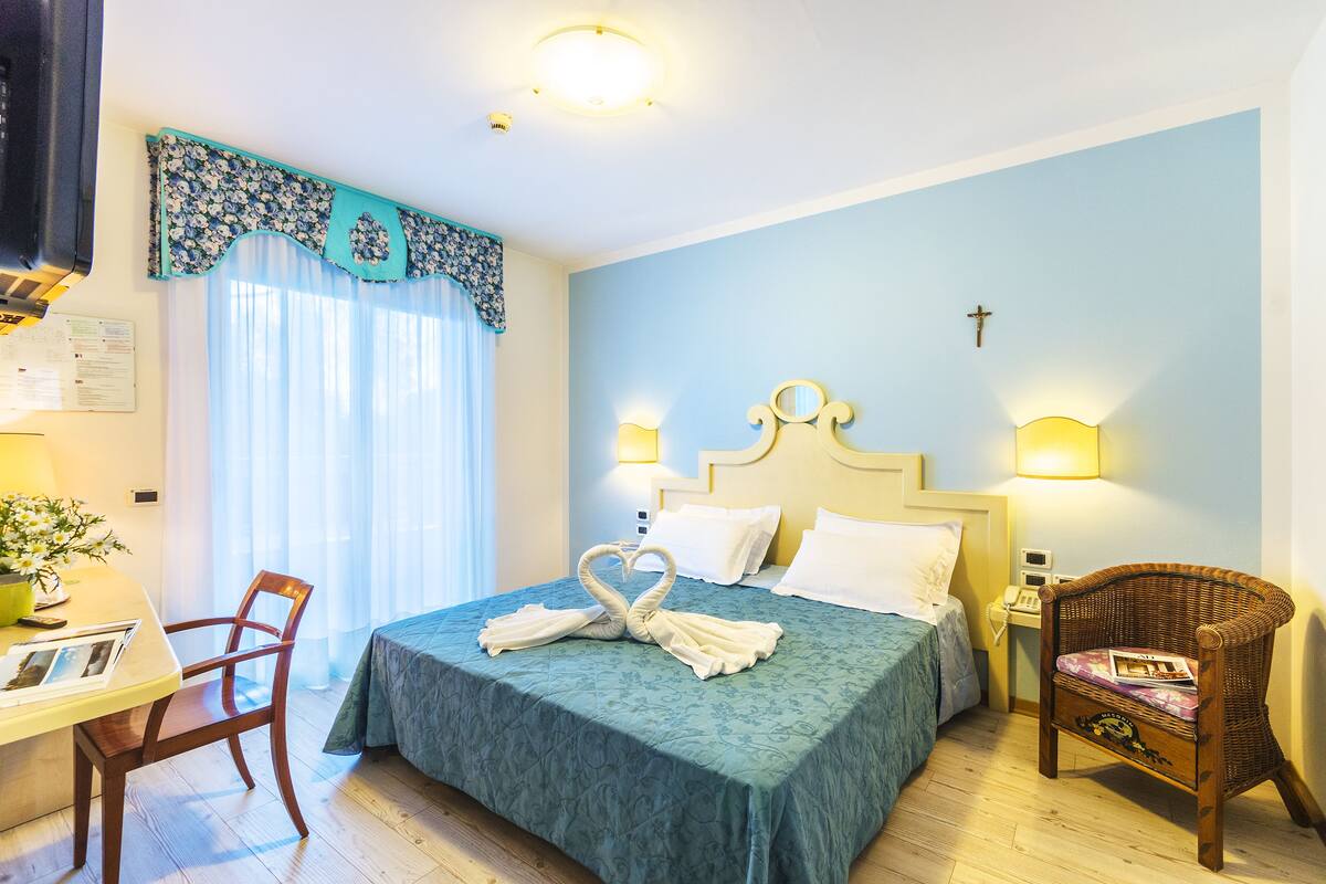 A neatly made bed with a blue bedspread, a chair, a desk with a plant, and a wall-mounted cross.