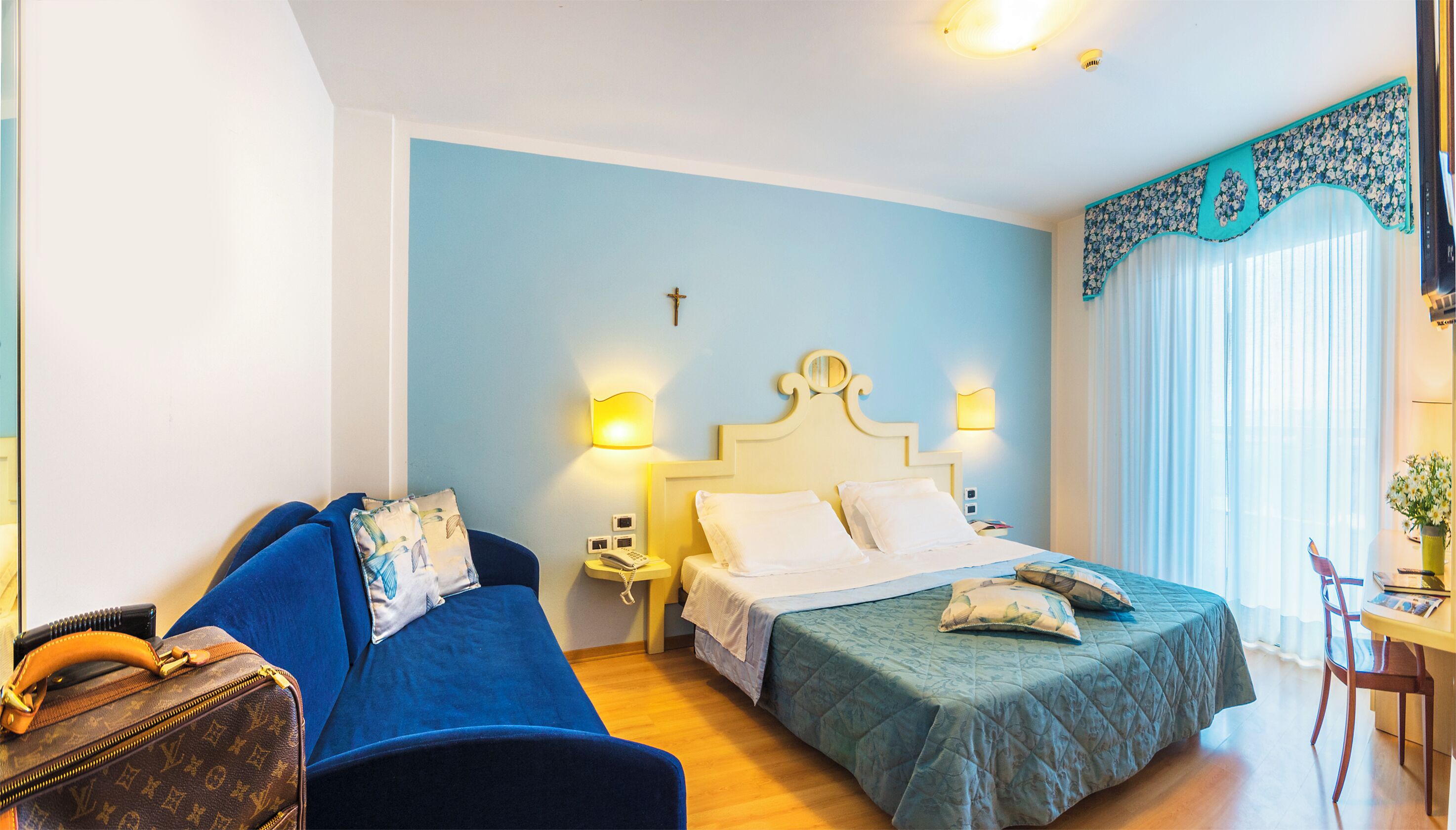 A hotel room with a bed, a blue sofa, a small table with a chair, and a window with curtains.