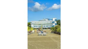 A hotel with a large building, multiple floors, and a parking area with several white benches. - Hotel Panoramic (Caorle)