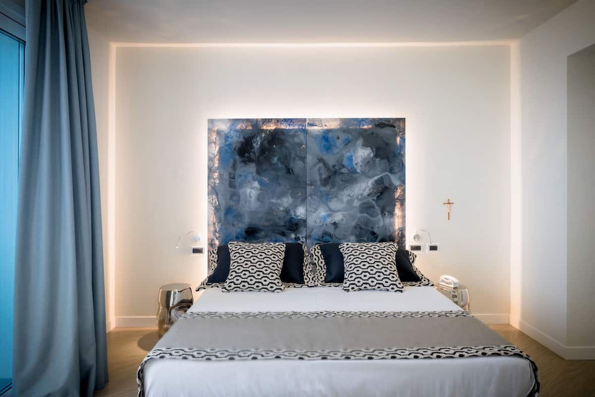 A modern bedroom with a large bed, a headboard with abstract artwork, bedside lamps, and a cross on the wall.